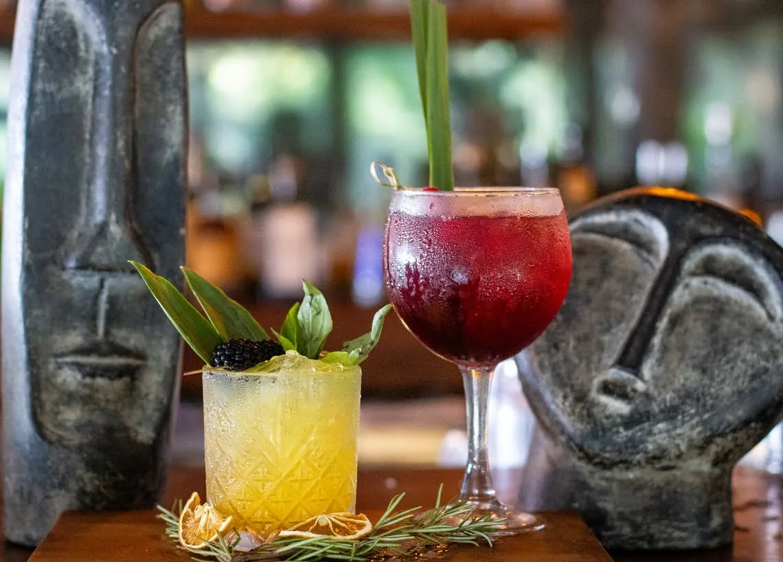 Two tropical cocktails and tiki statues at The Fives Corpo.