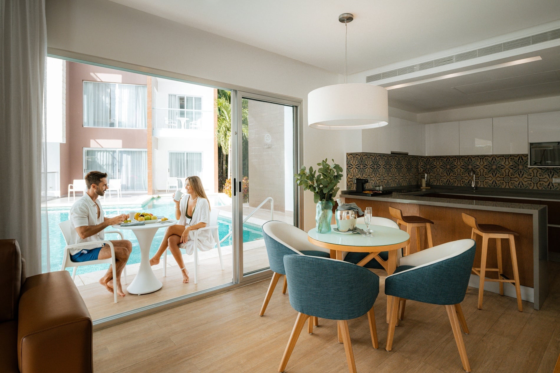 Spacious hotel suite, couple enjoying breakfast by pool, modern dining area and kitchen.