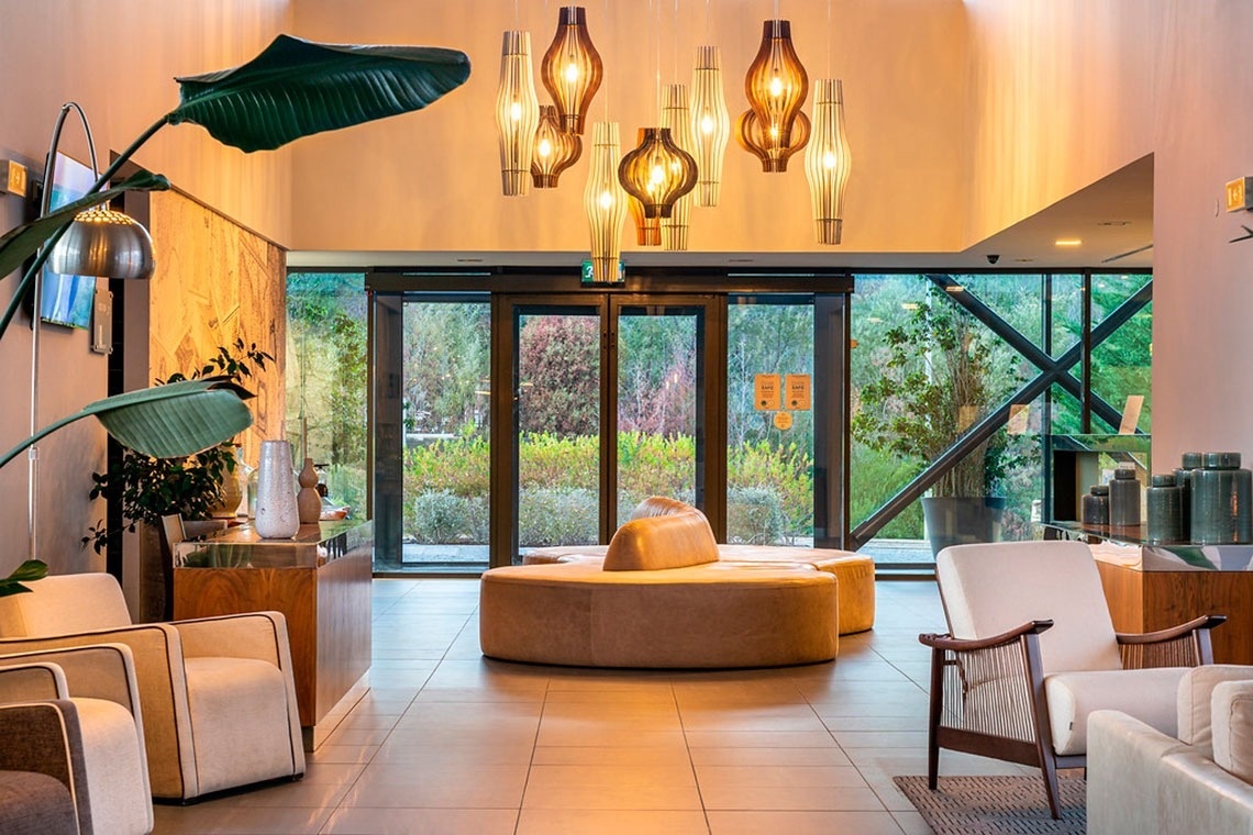 Lounge moderno com sofá curvo e poltronas, próximo às portas de vidro e à natureza. Monchique Resort & SPA por Flagworld Hotels.