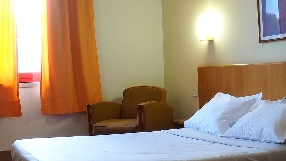 Quarto aconchegante com cama arrumada e poltrona. Flag Hotel Porto-Maia.
