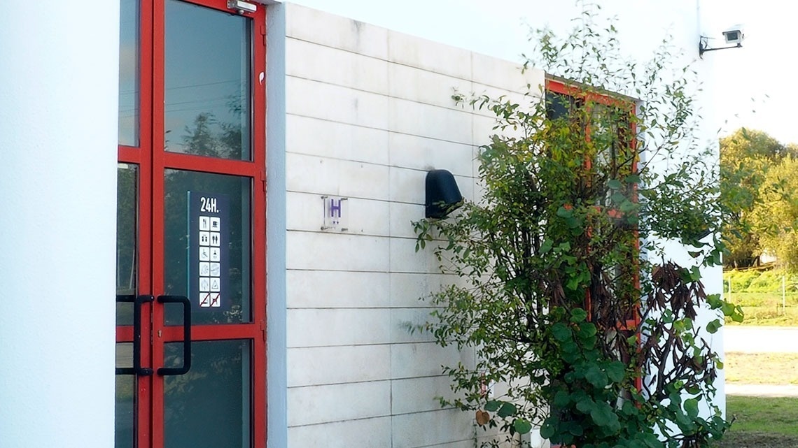 Fachada do Flag Hotel Porto-Maia com porta vermelha, sinais informativos e paisagem ao fundo.