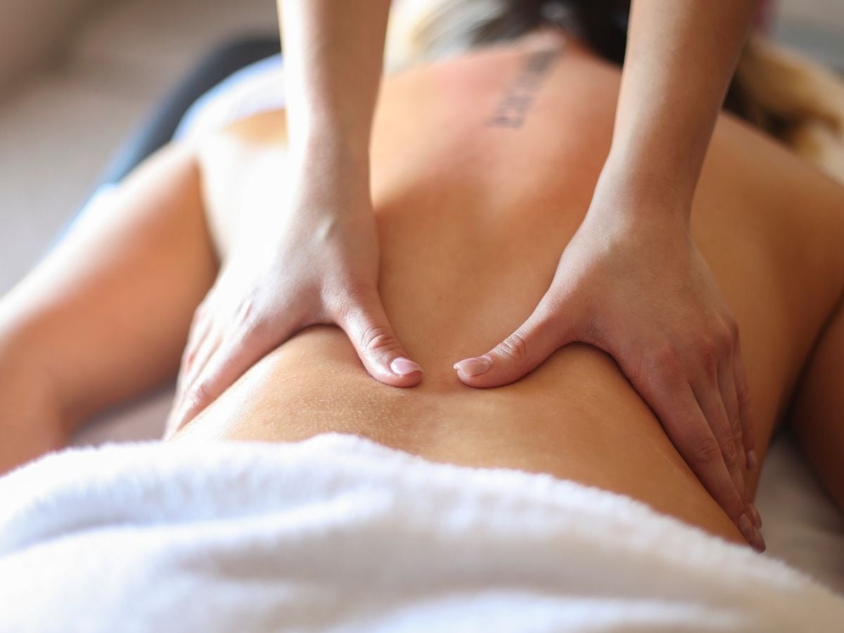 Uma pessoa está recebendo uma massagem nas costas com as mãos de um massagista, enquanto está deitada de bruços e parcialmente coberta por uma toalha branca.