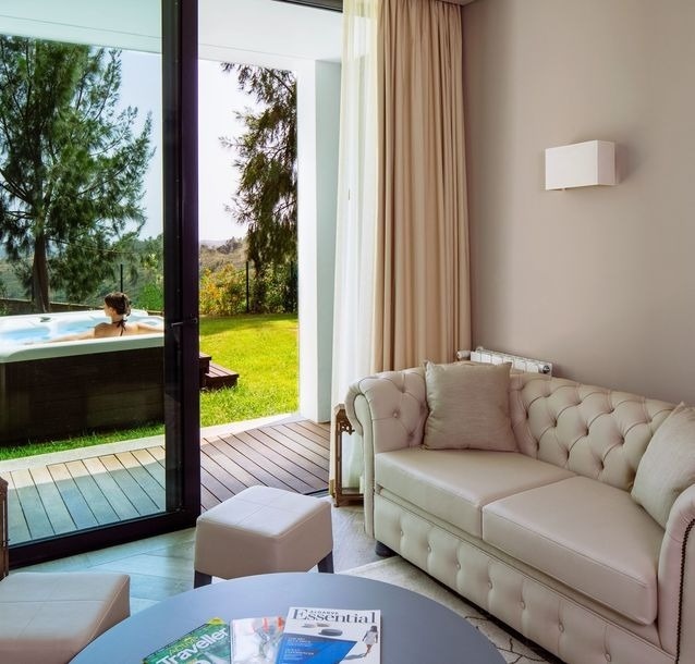 Uma mulher relaxa numa banheira de hidromassagem no jardim, visível através de uma grande porta de vidro de uma sala de estar com um sofá bege e uma mesa de centro.