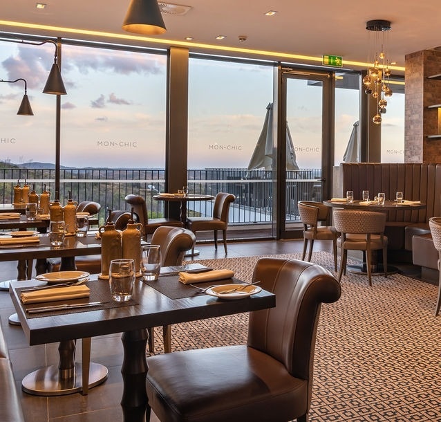 A modern restaurant interior features elegant brown leather seating, dark wooden tables set for dining, and large windows offering a panoramic view of a landscape under a cloudy sky.