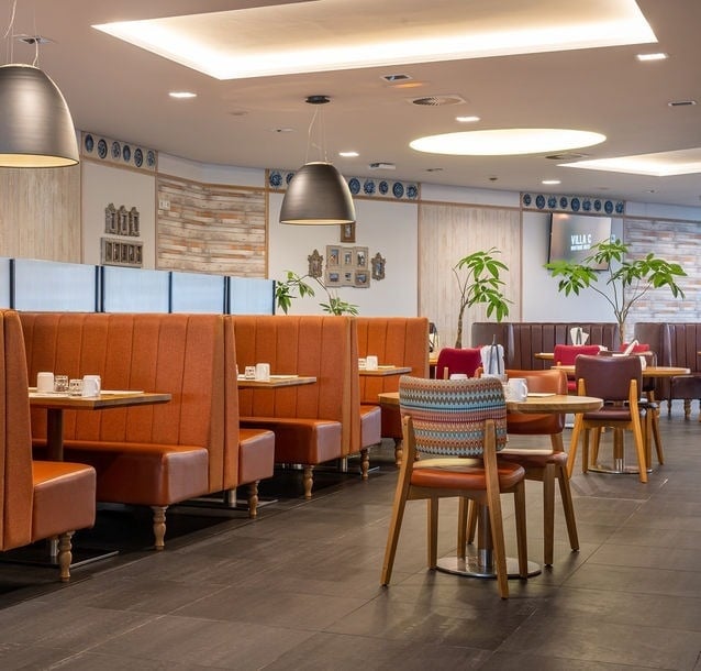 A modern restaurant dining room is depicted with rows of orange upholstered booths, wooden tables, and various chairs, illuminated by contemporary lighting fixtures and decorated with potted plants.