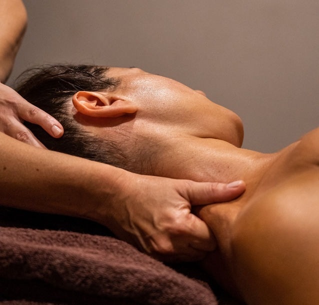Uma pessoa deitada de lado recebe uma massagem relaxante no pescoço e ombros, com as mãos do massagista visíveis aplicando pressão.