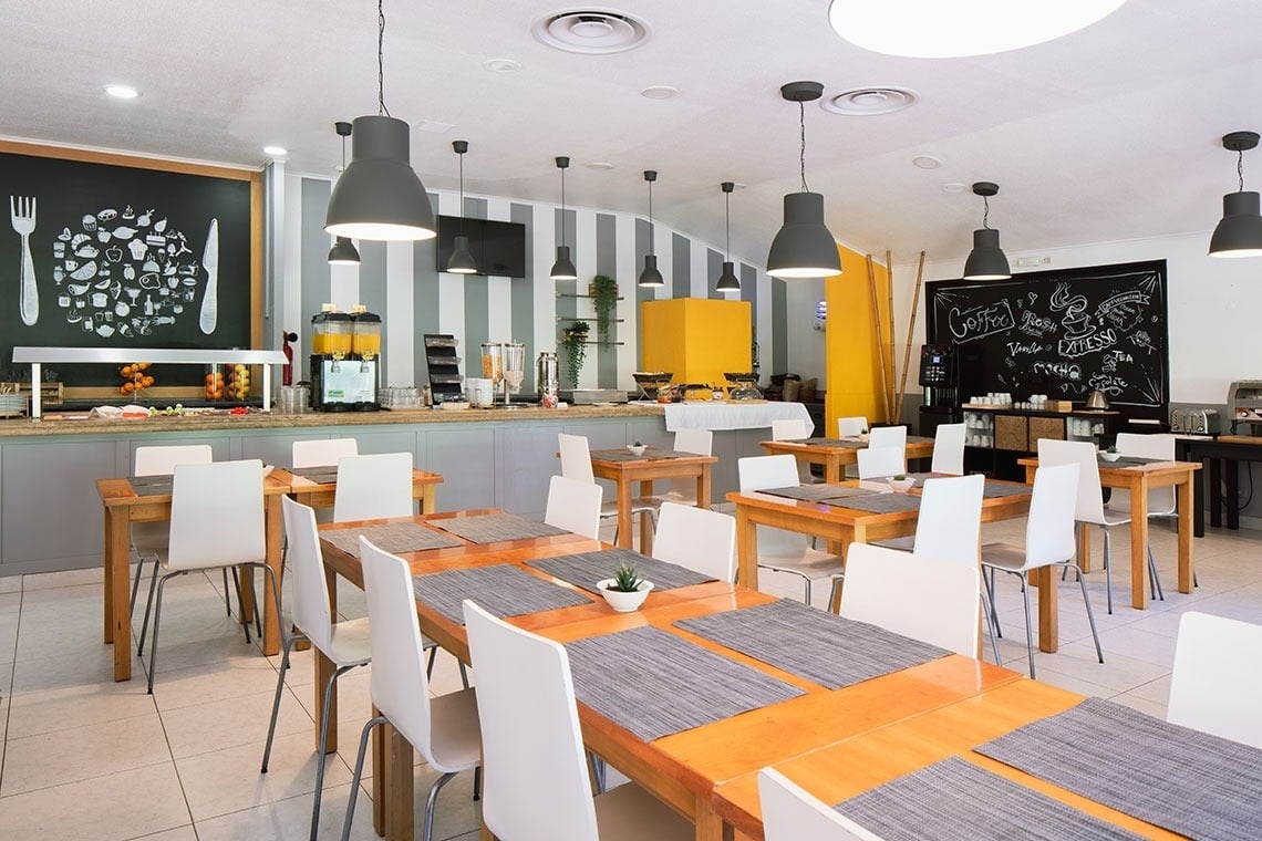 Buffet de café da manhã no Campanile Setúbal, com mesas de madeira e cadeiras brancas.