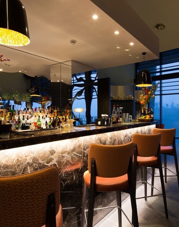 A luxurious bar interior features a marble counter with warm underlighting, comfortable orange bar stools, and a stocked liquor display, enhanced by ambient and pendant lighting.