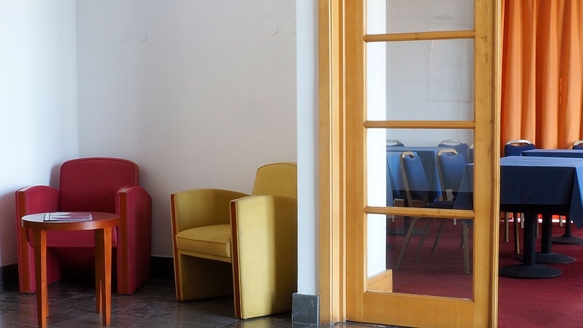 Sala de estar com poltronas vermelhas e amarelas, mesa de centro e sala de conferências ao fundo. Flag Hotel Porto-Maia.