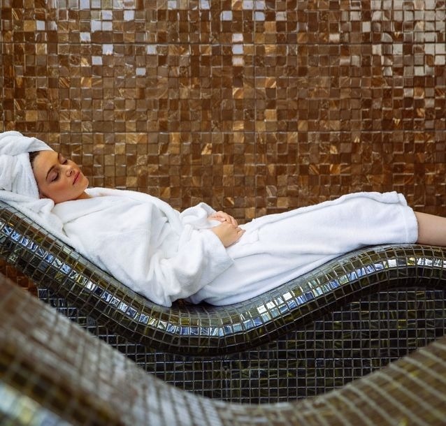 Uma mulher de roupão branco e toalha na cabeça está deitada e relaxando em uma espreguiçadeira de mosaico em um ambiente de spa.