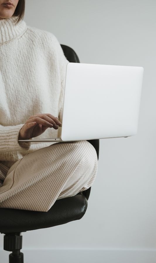 una mujer con un suéter blanco está sentada en una silla usando una computadora portátil