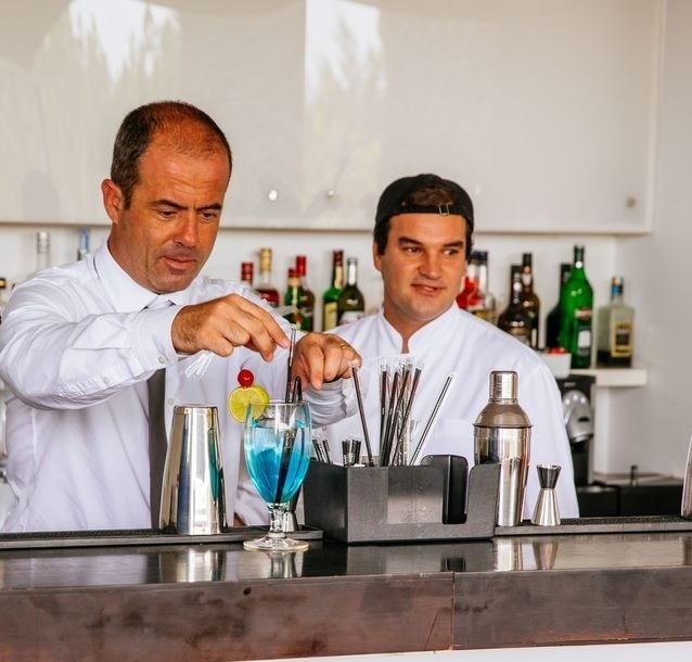 Two bartenders are preparing a blue cocktail at a bar, with one garnishing the drink and the other observing.