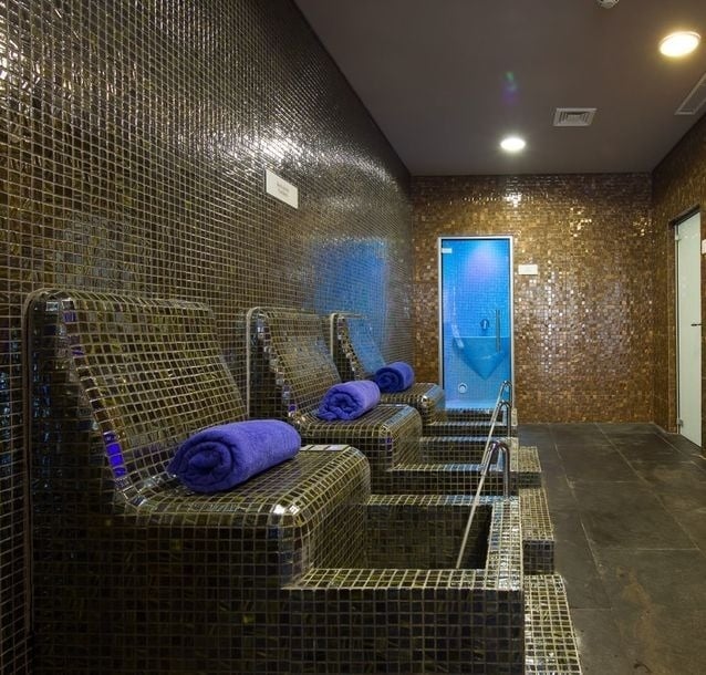 Esta imagem exibe uma sala de spa com cadeiras reclináveis e paredes revestidas de mosaicos dourados e refletivos, com toalhas roxas sobre as cadeiras e uma porta azul iluminada ao fundo.