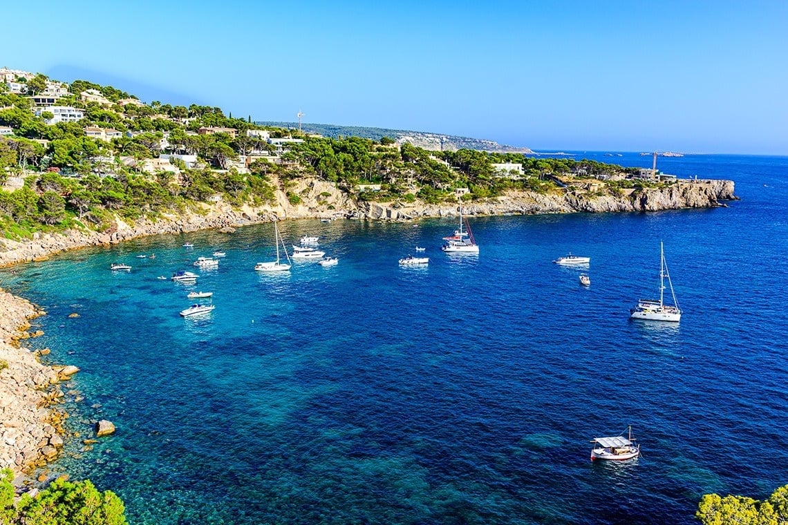 Numerous boats are anchored in a clear blue bay, bordered by a lush, residential hillside under a bright sky.