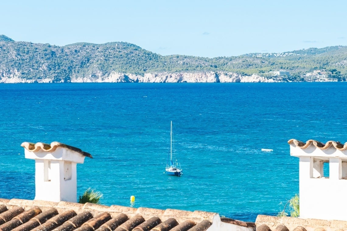 A scenic view features a blue sailboat anchored in the turquoise sea, framed by a foreground of terracotta rooftops and white chimneys, with a rugged, tree-covered coastline and distant mountains under a clear sky.