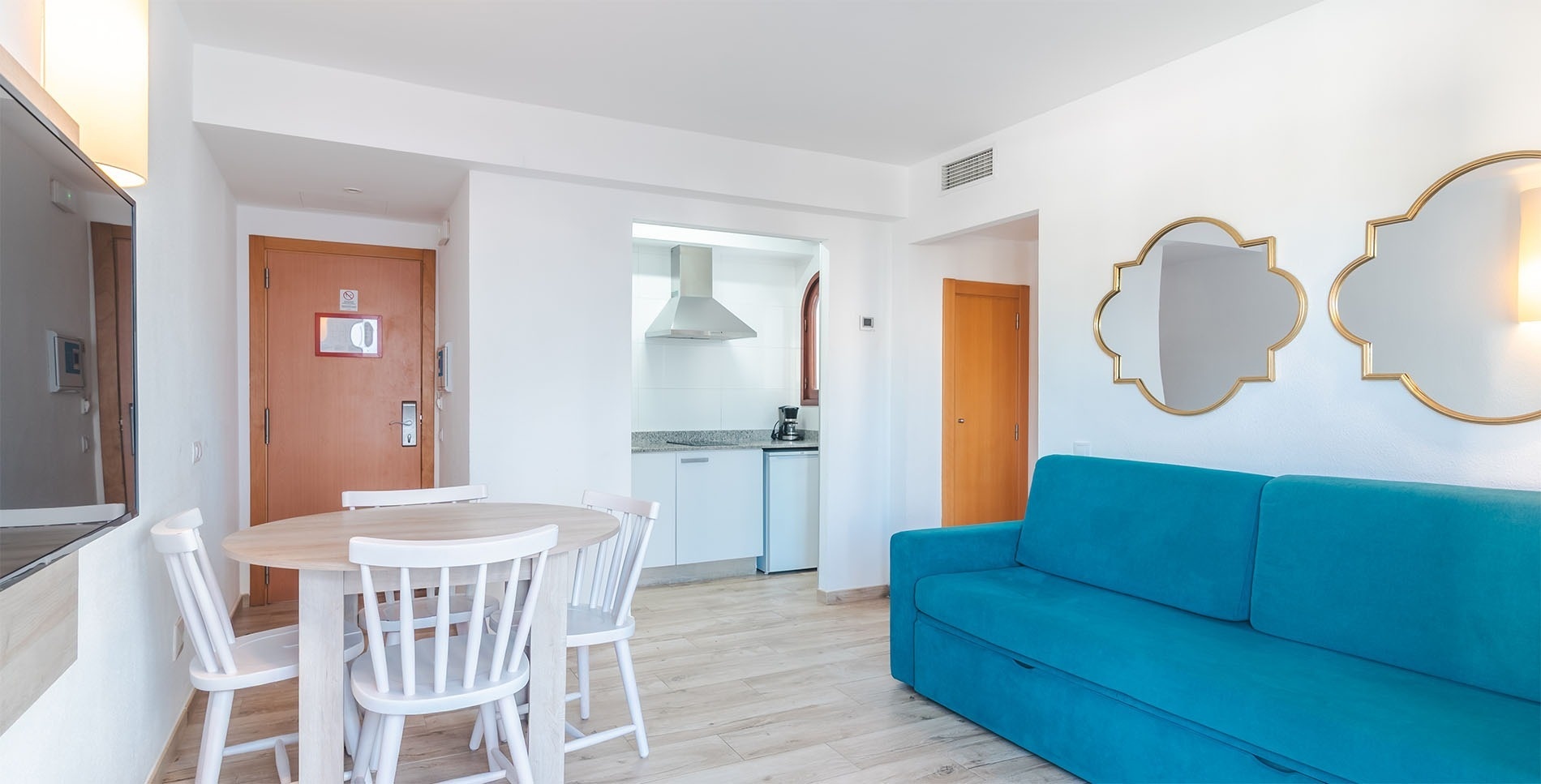 Un luminoso y moderno apartamento tipo estudio con un sofá azul, una mesa de comedor de madera clara con sillas blancas y una cocina compacta integrada al fondo.