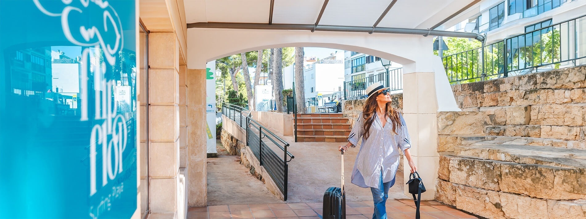Una mujer con sombrero y equipaje camina por un pasillo exterior de baldosas, junto a un edificio con un llamativo letrero azul y bajo una arcada.