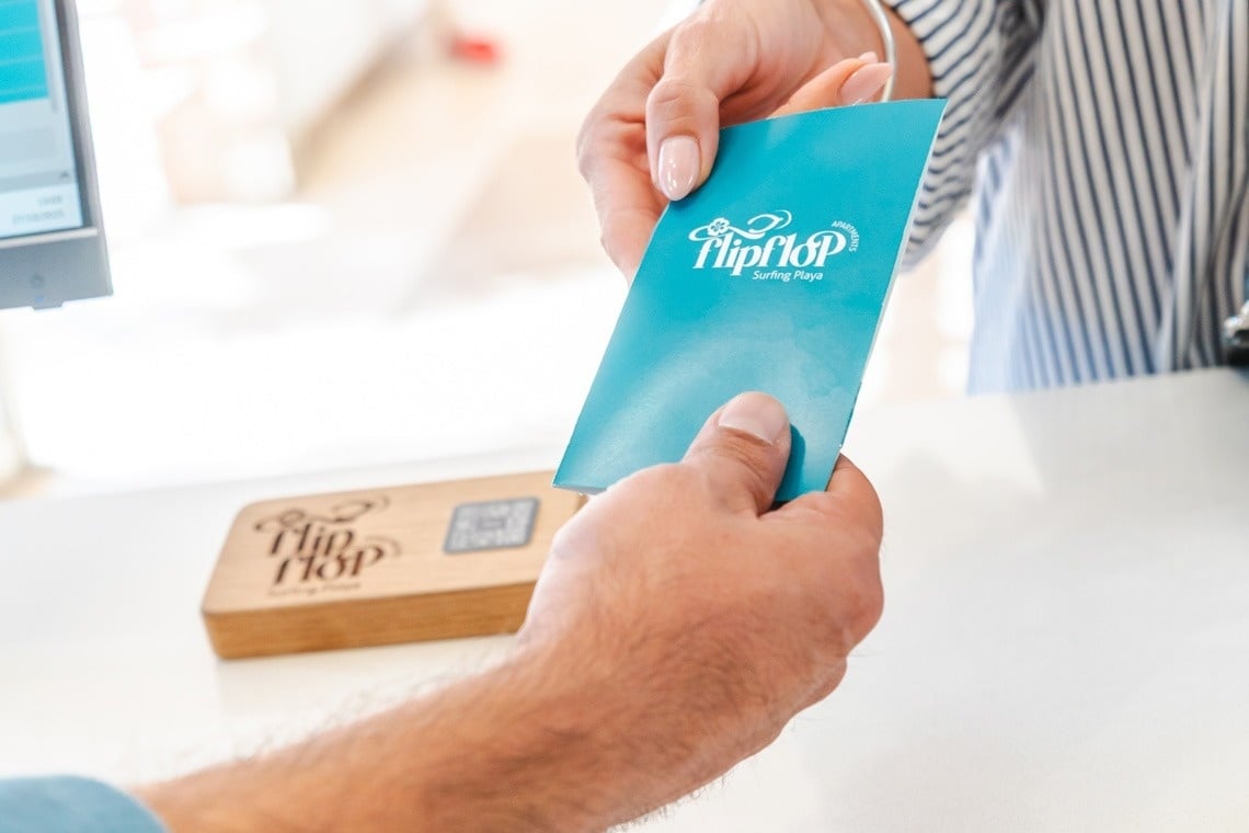 Dos pares de manos intercambian una tarjeta azul con el logo de "FlipFlop Surfing Playa".
