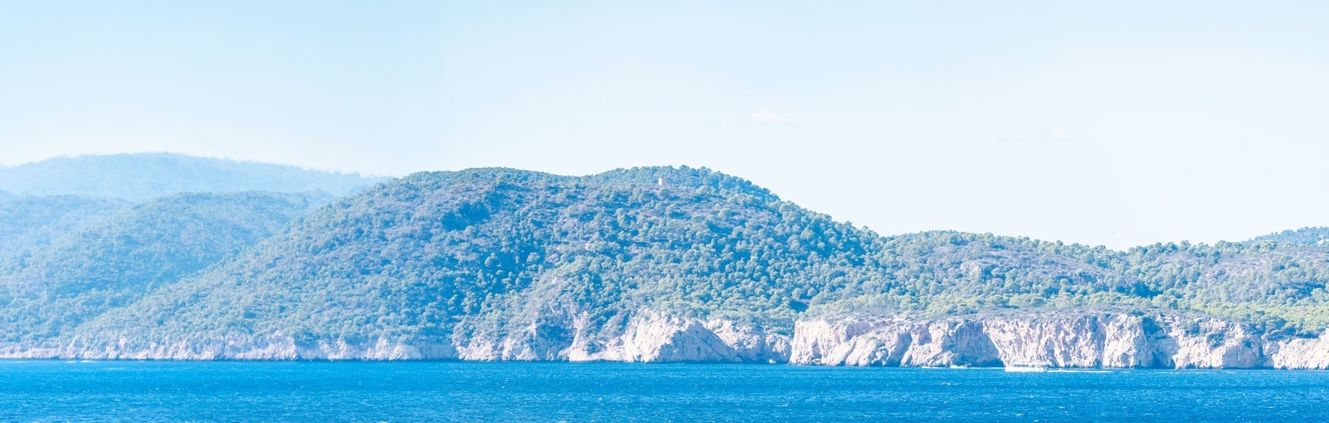 A scenic view features a vibrant blue sea with a sailboat, backed by tree-covered hills and rocky cliffs, framed by green foliage on the left.