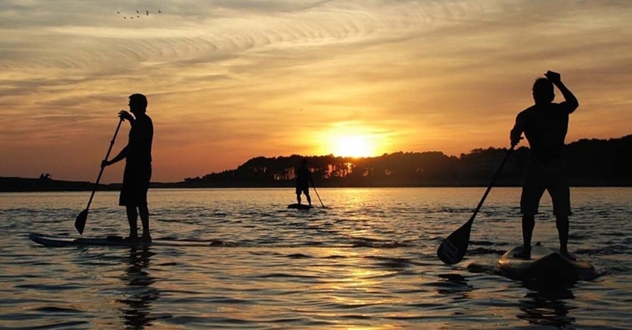 três pessoas estão remando pranchas de remo na água ao pôr do sol