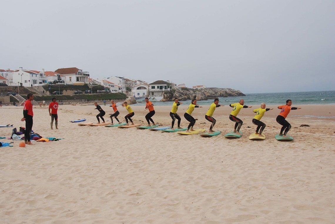 Um grupo de alunos de surf pratica posições em pranchas na areia da praia, sob a supervisão de instrutores, com uma vila costeira ao fundo.