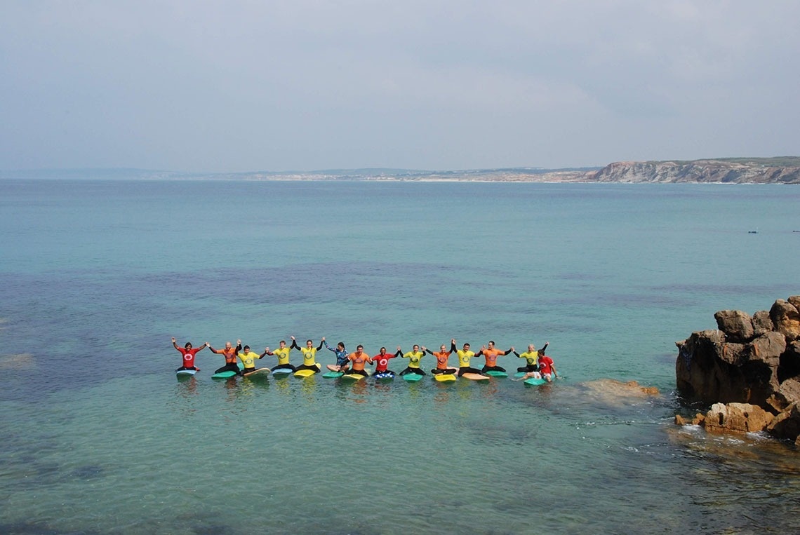 Um grupo de pessoas está sentado em pranchas de surf, de mãos dadas em fila na água azul-turquesa do mar, com uma costa rochosa e falésias ao fundo.