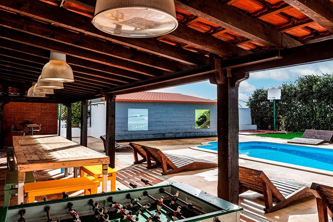 Uma área de lazer externa coberta, com teto de madeira, mesa de pebolim, mesa comprida e luminárias, oferece vista para uma piscina com espreguiçadeiras e um edifício ao fundo.