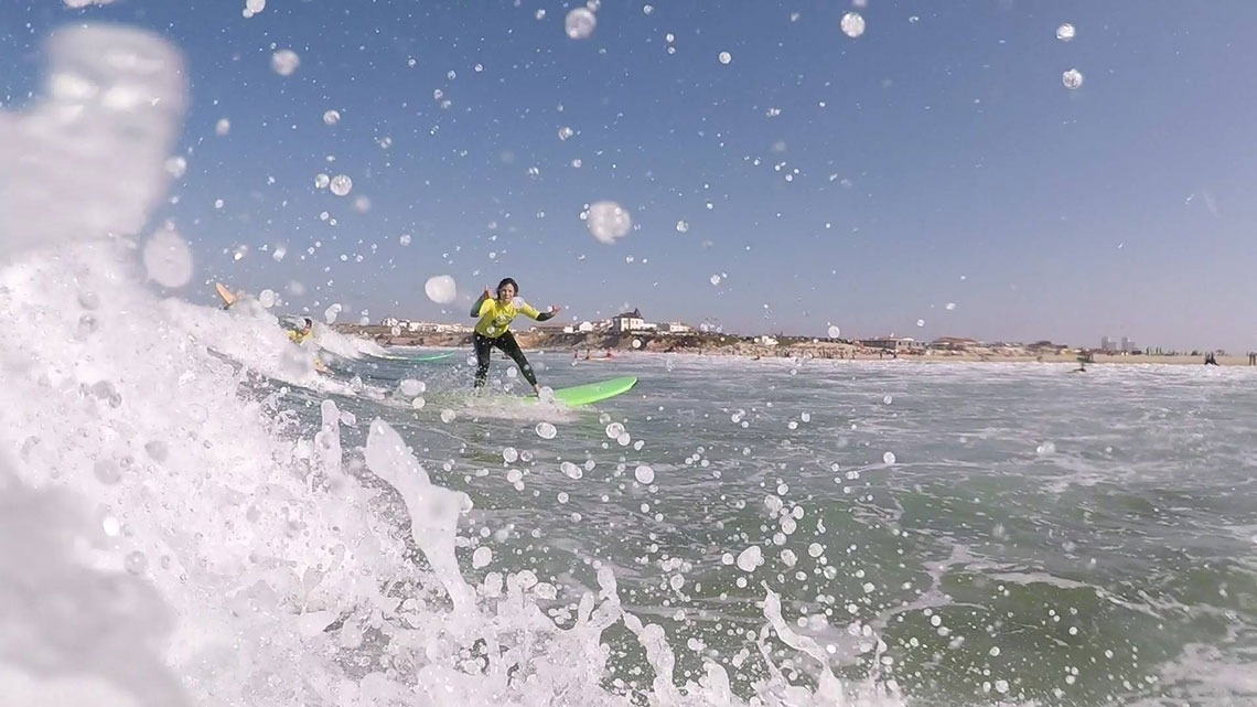 Uma surfista em uma prancha verde é vista a cavalgar uma onda no oceano, cercada por salpicos de água, com uma paisagem costeira e edifícios ao fundo sob um céu azul.