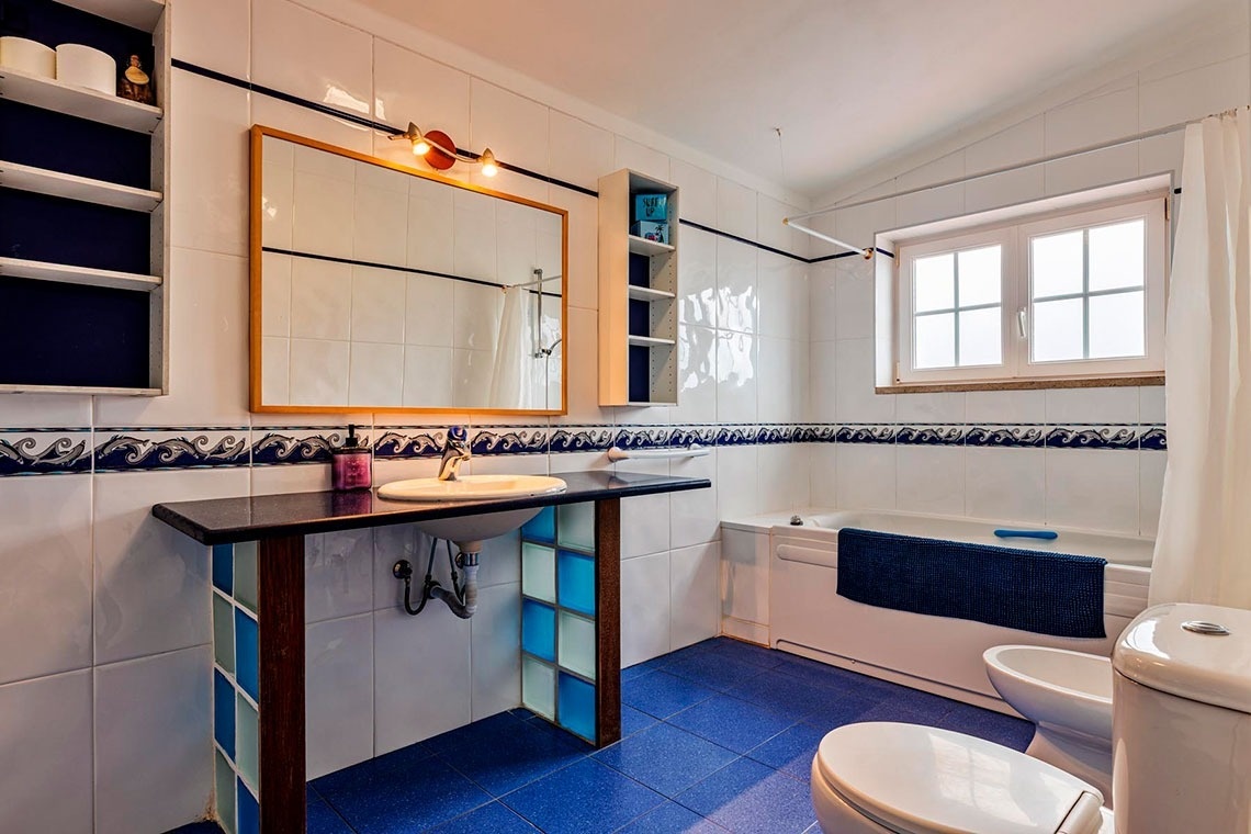 Um banheiro com azulejos brancos e azuis, lavatório com bancada escura, espelho, banheira e louças sanitárias.