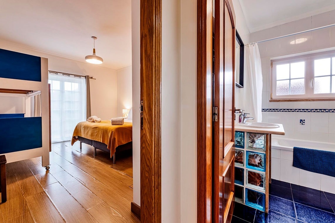 A imagem apresenta uma vista dividida entre um quarto com piso de madeira e beliches à esquerda, e uma casa de banho com azulejos azuis e brancos, banheira e um lavatório com base de blocos de vidro à direita.