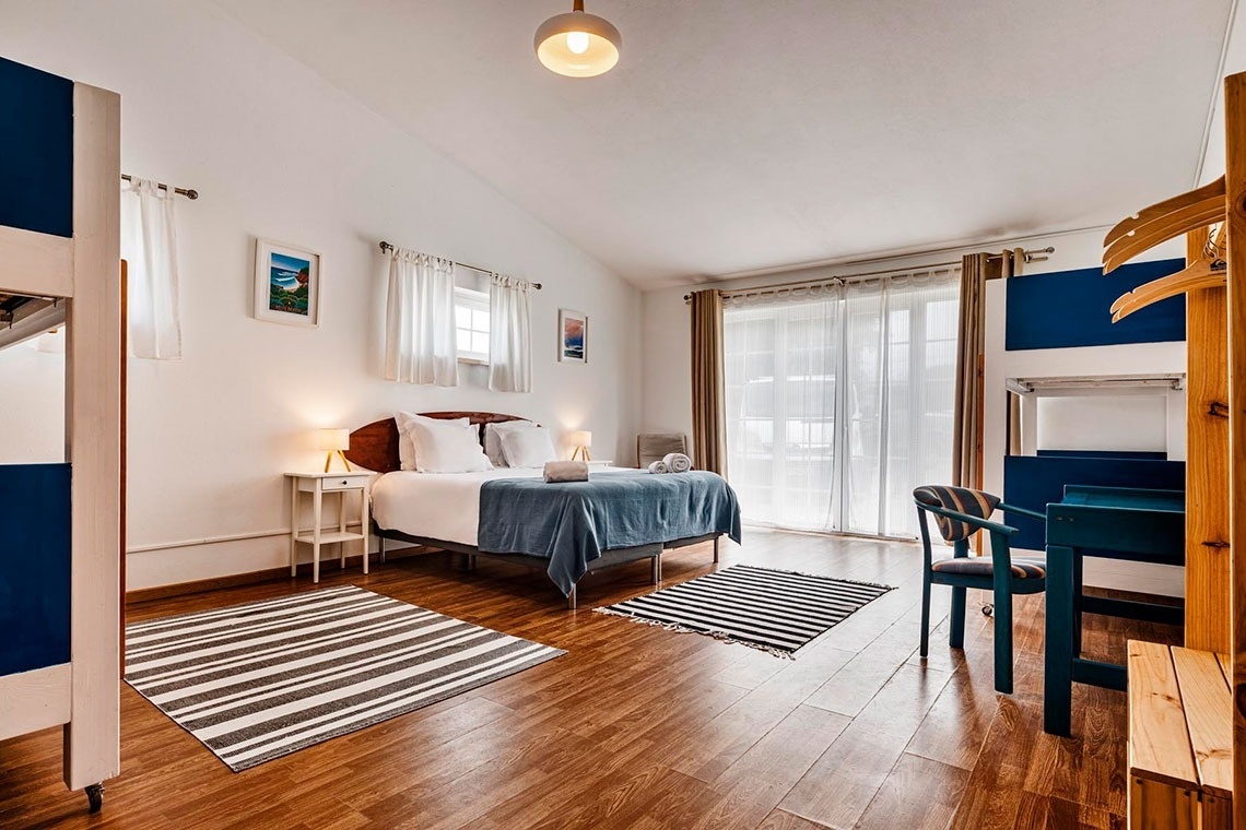 Um quarto luminoso e moderno, com uma cama de casal, tapetes listrados no chão de madeira e detalhes em azul.