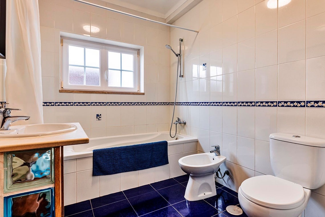 Uma casa de banho com paredes de azulejos brancos decorados com uma faixa azul, chão de azulejos azuis, banheira, bidé, sanita, lavatório e uma janela.