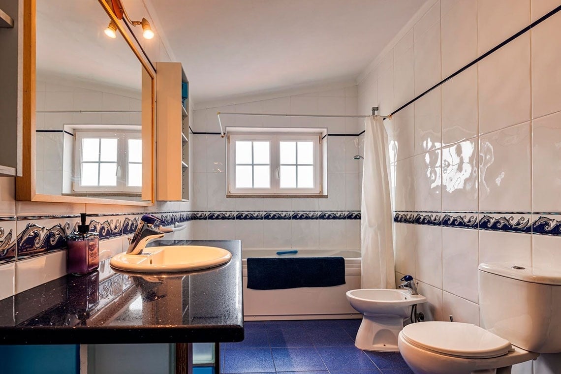 Uma casa de banho funcional e luminosa, com paredes em azulejo branco e frisos azuis, chão azul, e equipada com lavatório, espelho, banheira, bidé e sanita.