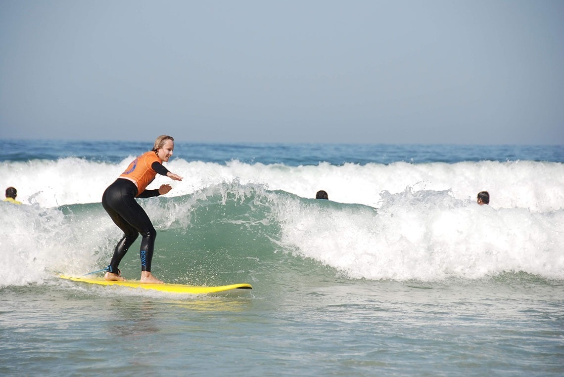 Uma mulher surfista com uma roupa de mergulho preta e laranja cavalga uma onda em uma prancha amarela, com outras pessoas na água ao fundo.