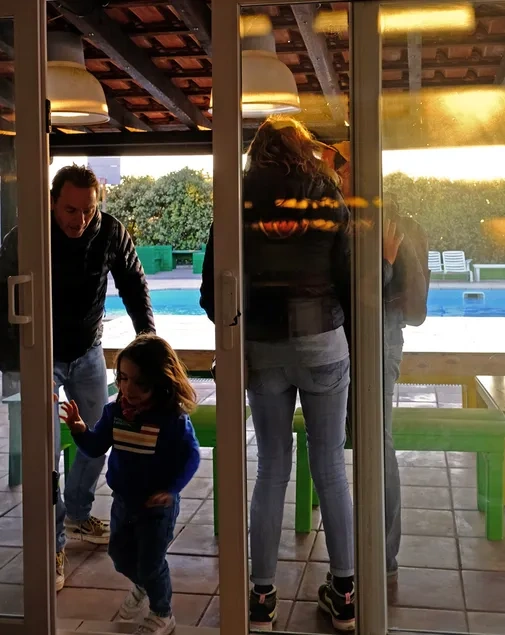 Pessoas, incluindo um homem e uma criança, são vistas através de uma porta de vidro com reflexos, com uma piscina e vegetação ao fundo.