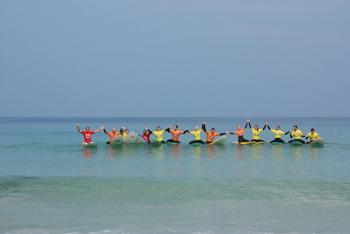 Um grupo de pessoas sentadas em pranchas de surfe no mar, de mãos dadas e com os braços levantados.