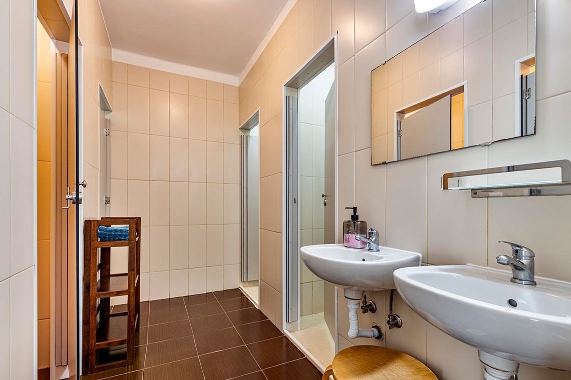 Um banheiro com paredes e chão de azulejos claros e escuros, apresentando dois lavatórios, um espelho e várias portas para chuveiros ou cabines.