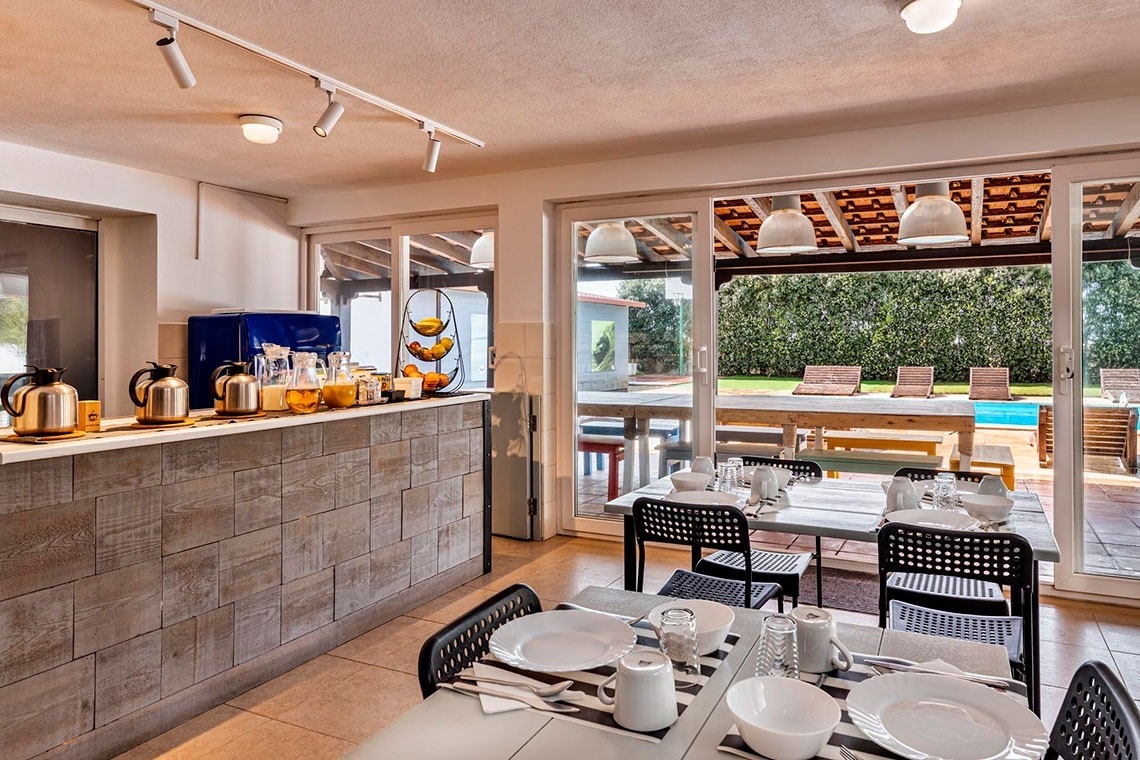 Uma área de refeições luminosa, com um balcão de pequeno-almoço e mesas postas, que se abre para um espaço exterior com piscina e espreguiçadeiras.