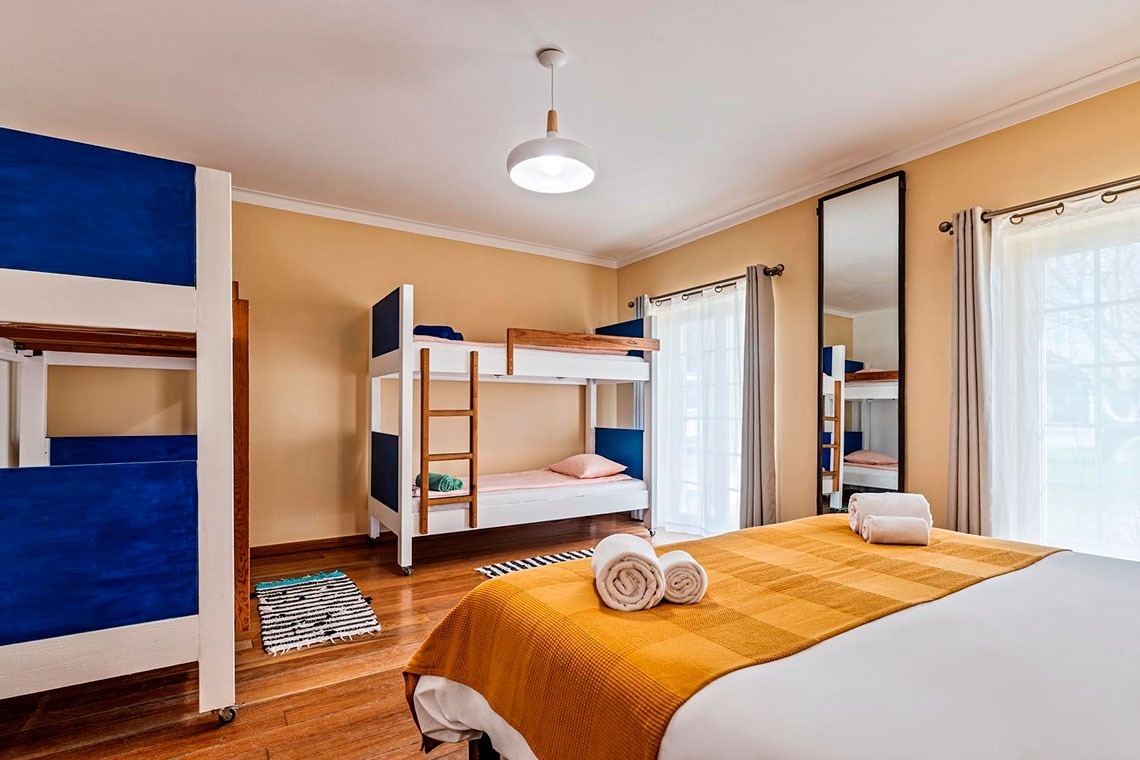 Um quarto de tons pêssego é apresentado com várias opções de dormir, incluindo beliches de cores azul e branco e uma cama de casal com um cobertor amarelo e toalhas enroladas, ao lado de um espelho de corpo inteiro e janelas com cortinas.