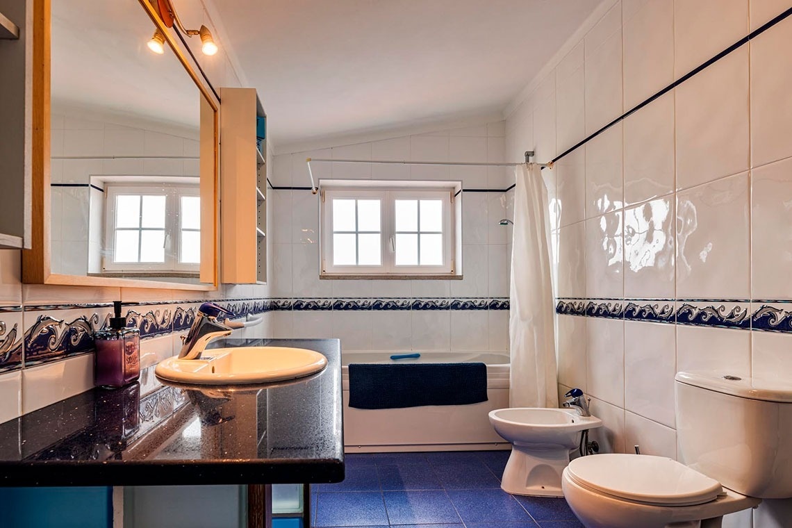 Uma casa de banho bem iluminada, com paredes de azulejo branco decoradas com uma faixa azul e chão de azulejo azul, equipada com lavatório, espelho, janela, banheira, bidé e sanita.