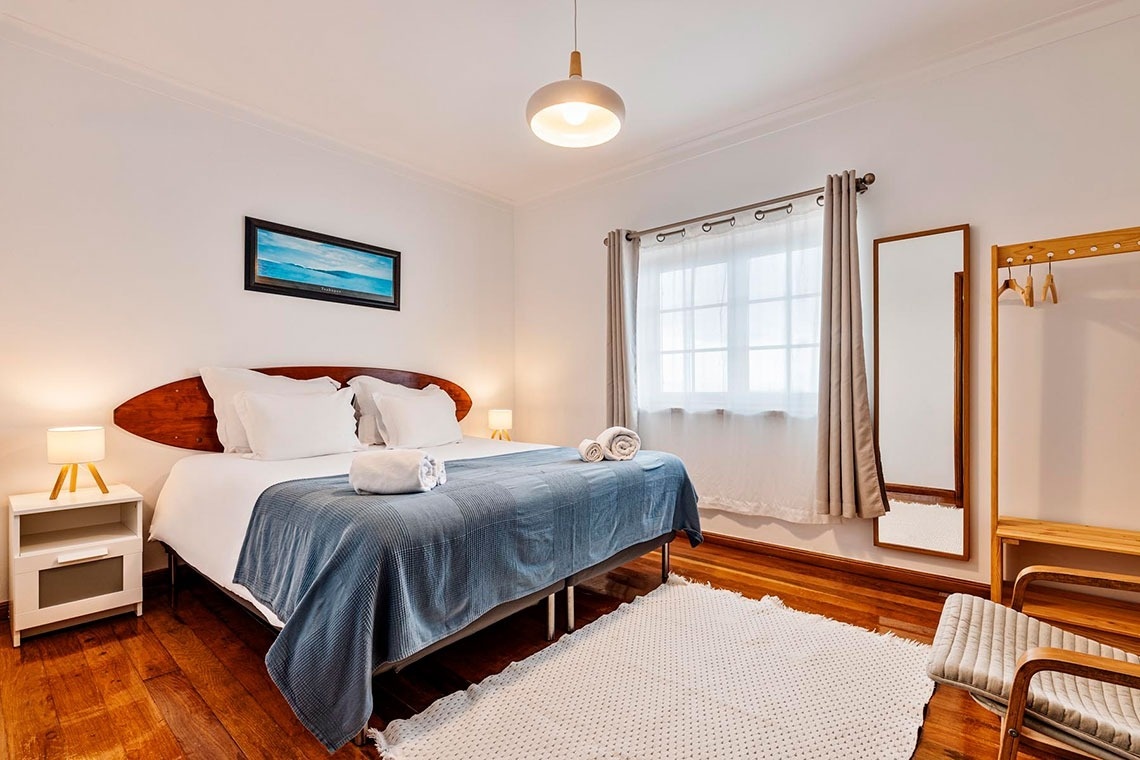 Um quarto luminoso com uma cama de casal, cabeceira de madeira estilo prancha de surf, mesas de cabeceira com candeeiros, uma janela com cortinas e piso de madeira com um tapete.