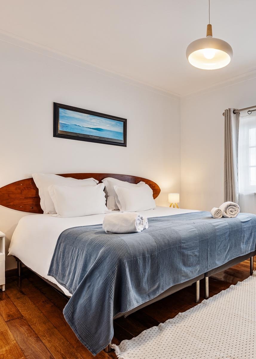 Um quarto luminoso apresenta uma cama com cabeceira de madeira que lembra uma prancha de surf, roupa de cama branca com um cobertor azul e um quadro de paisagem marinha pendurado na parede acima dela.