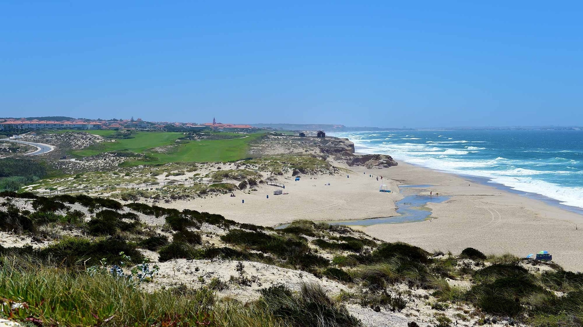 Uma paisagem costeira panorâmica com uma praia extensa e ondas azuis, dunas arenosas, um campo de golfe verdejante e edifícios com telhados de terracota à distância sob um céu límpido.