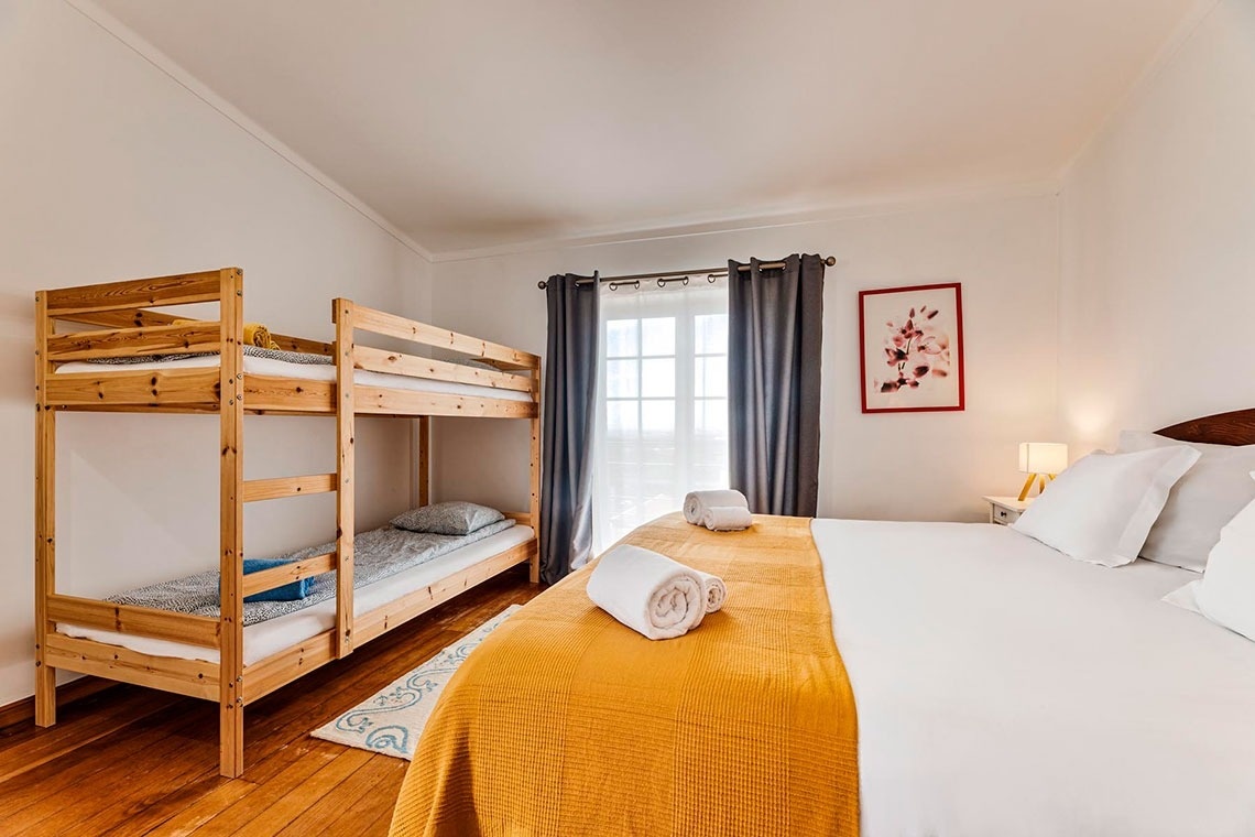 Um quarto de dormir luminoso com beliches de madeira, uma cama de casal com edredom amarelo e branco, cortinas cinzas e um quadro floral.