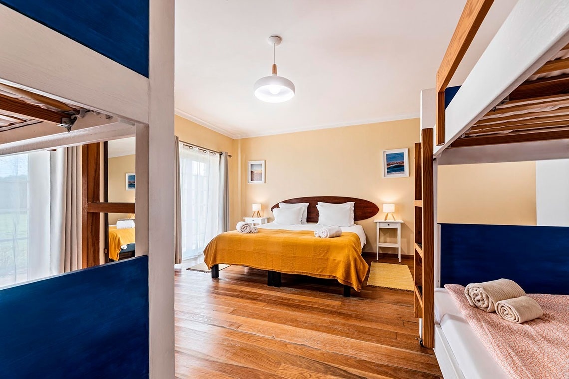 Um quarto de dormir luminoso com paredes amarelas e piso de madeira apresenta uma cama de casal central com colcha amarela, ladeada por mesas de cabeceira, e um beliche parcialmente visível em ambos os lados.