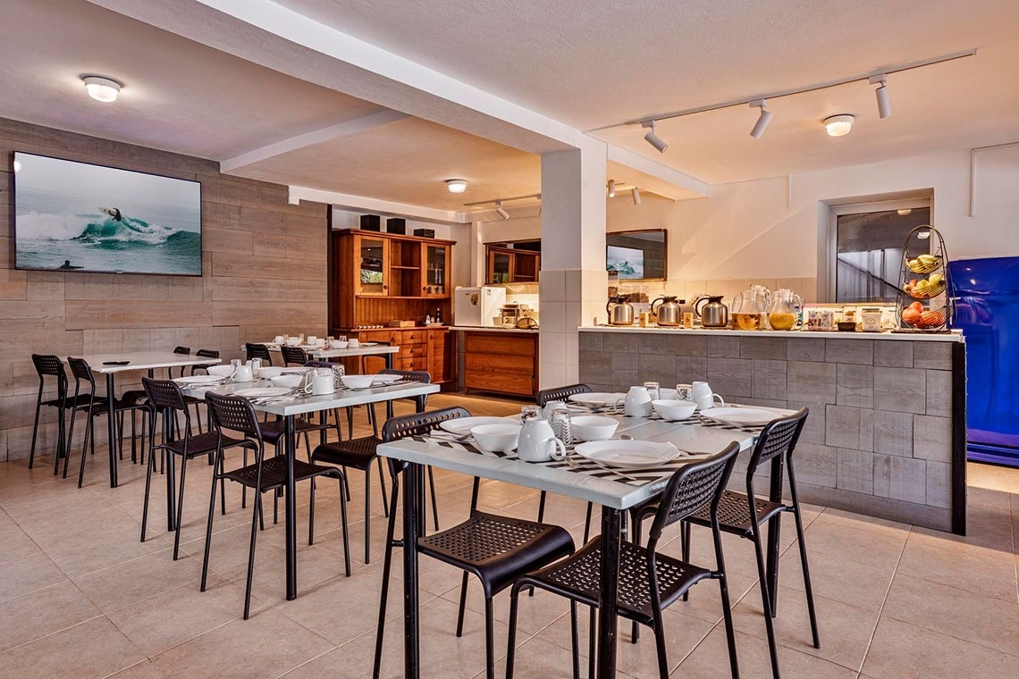 Uma sala de jantar ou café da manhã com várias mesas prontas, cadeiras pretas, uma televisão, e uma área de buffet com bebidas e frutas.