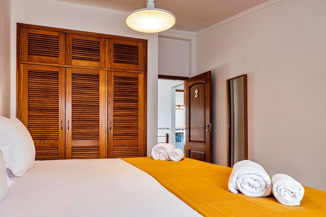 Um quarto bem iluminado apresenta uma cama arrumada com um cobertor amarelo, um guarda-roupa de madeira e uma porta entreaberta com o número 2.