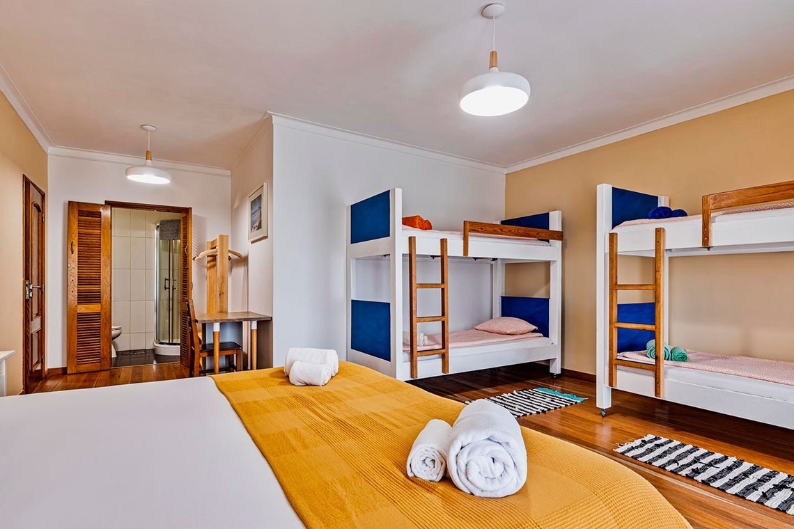 Um quarto espaçoso com uma cama de casal em primeiro plano e várias beliches ao fundo, com acesso a um banheiro.