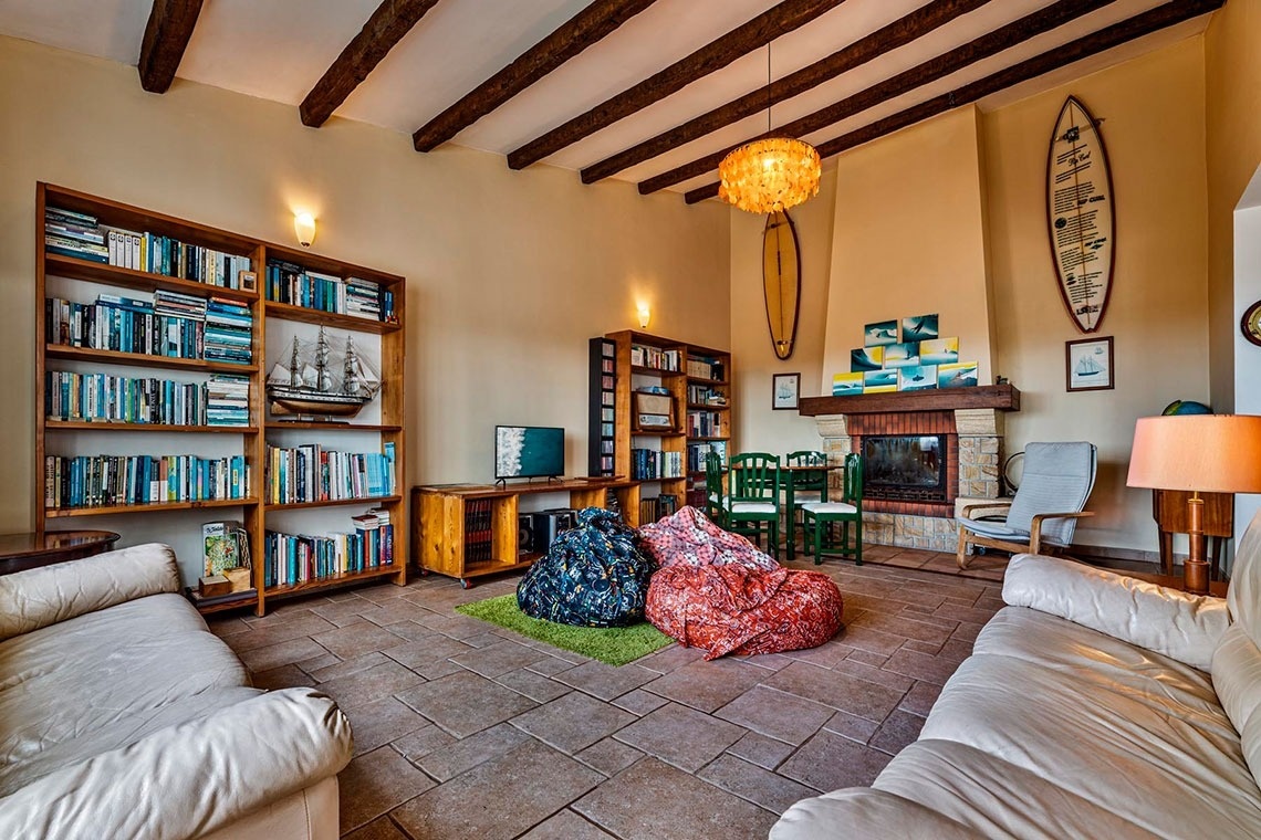 Uma sala de estar rústica e acolhedora, com estantes de livros, uma lareira de pedra, vigas de madeira expostas no teto, puffs coloridos no chão e elementos de decoração de surf e náuticos.