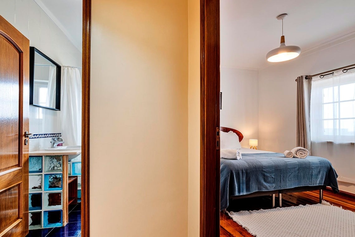 Duas portas abertas revelam um banheiro com pia à esquerda e um quarto com cama à direita.